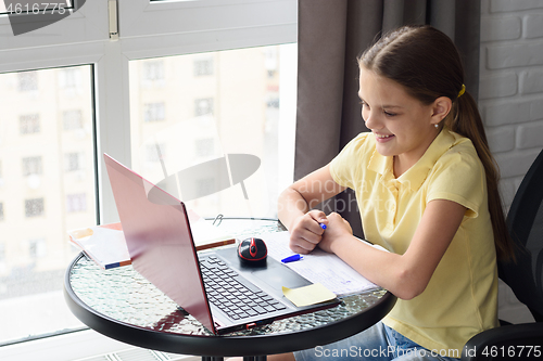 Image of Girl joyfully looks at the laptop screen while sitting at a table in the apartment and doing homework