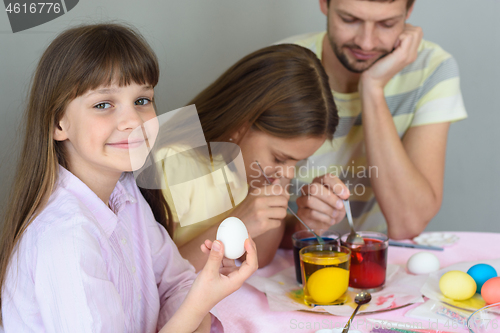 Image of A girl with an egg in her hands looked into the frame, in the background the family paints eggs in glasses with dyes