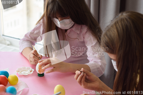 Image of Two girls in medical masks paint eggs with a brush while preparing for Easter