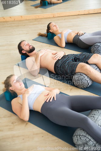 Image of Restorative yoga with a bolster. Group of young sporty attractive people in yoga studio, lying on bolster cushion, stretching and relaxing during restorative yoga. Healthy active lifestyle