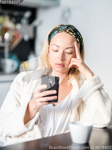 Image of Beautiful caucasian woman at home, feeling comfortable wearing white bathrobe, taking some time to herself, drinking morning coffee and reading news on mobile phone device in the morning