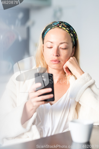 Image of Beautiful caucasian woman at home, feeling comfortable wearing white bathrobe, taking some time to herself, drinking morning coffee and reading news on mobile phone device in the morning