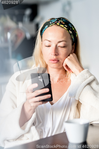 Image of Beautiful caucasian woman at home, feeling comfortable wearing white bathrobe, taking some time to herself, drinking morning coffee and reading news on mobile phone device in the morning