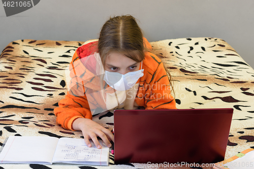 Image of A girl in a medical mask in pajamas lies on the bed and remotely learns via the Internet