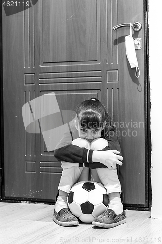 Image of A girl sits at the front door with a ball, but she is not allowed to go outside during quarantine