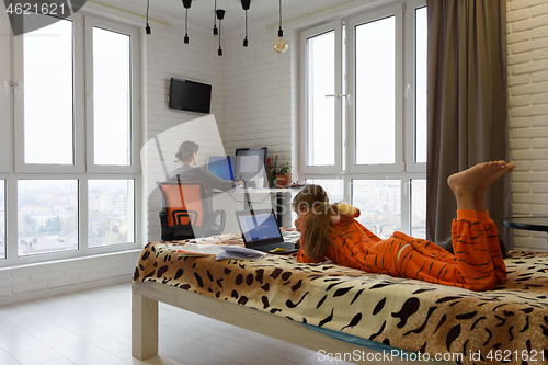 Image of The family is at home, the child lies on the bed and studies in a laptop, the mother in the background works remotely in the computer