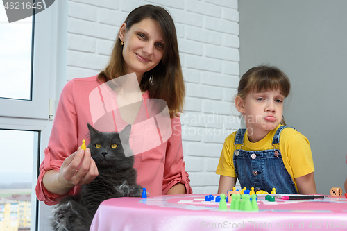 Image of At the table are a cheerful girl, a displeased girl and a domestic cat