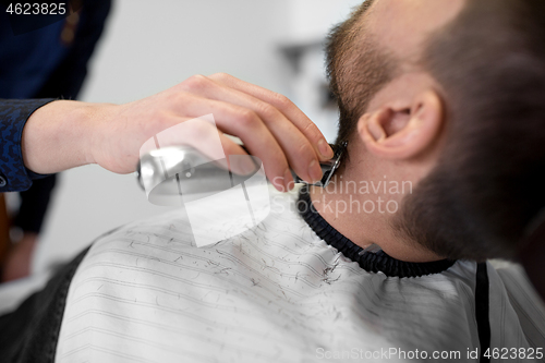 Image of man and barber with trimmer cutting beard at salon