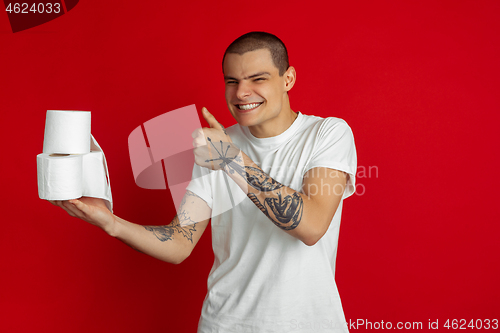 Image of Caucasian young man\'s portrait on red studio background - holding toilet papers, essential goods during quarantine and self-insulation