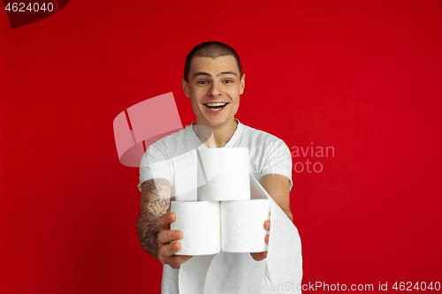 Image of Caucasian young man\'s portrait on red studio background - holding toilet papers, essential goods during quarantine and self-insulation