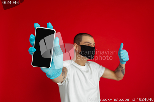 Image of Caucasian young man in protective face mask and medical gloves on red studio background - showing blank phone\'s screen