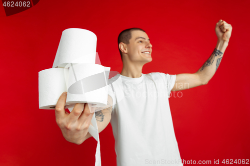 Image of Caucasian young man\'s portrait on red studio background - holding toilet papers, essential goods during quarantine and self-insulation