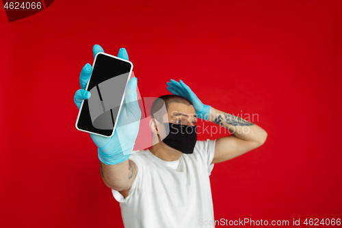 Image of Caucasian young man in protective face mask and medical gloves on red studio background - showing blank phone\'s screen