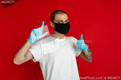 Image of Caucasian young man in protective face mask and medical gloves on red studio background - pointing and showing thumb up