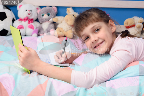 Image of A schoolgirl studies remotely at home, lies in bed and watches a video lesson