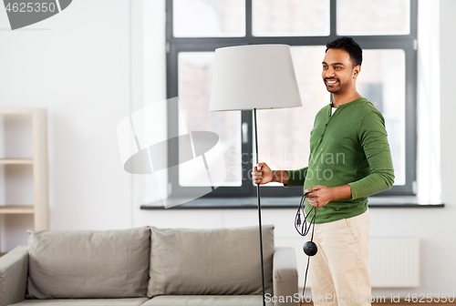 Image of happy indian man holding floor lamp at home