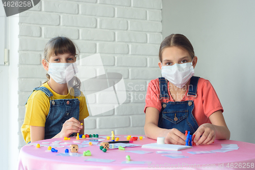 Image of Two girls in protective medical masks play board games at home