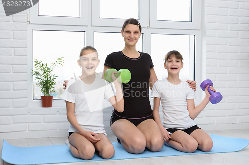 Image of Two cheerful girls and their mother sit on the floor with dumbbe