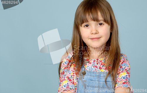 Image of Sweet little girl 6-7 years old posing in dungarees jeans and flower pattern blouse on blue