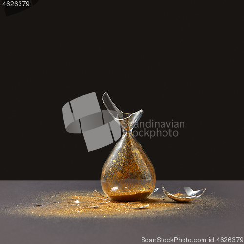 Image of Brocken vintage sandglass with golden sand on a black duotone background.