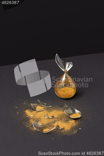 Image of Crashed outdated sandglass with golden sand on a black duotone background.