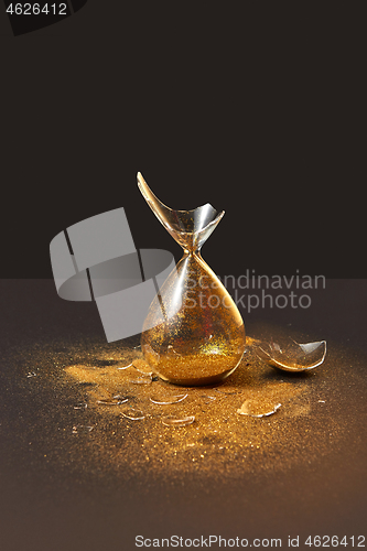 Image of Cracked antique hourglass with golden sand on a black duotone background.