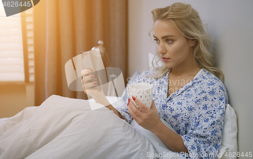 Image of Beautiful young woman browsing smartphone and holding mug of hot beverage sitting on bed