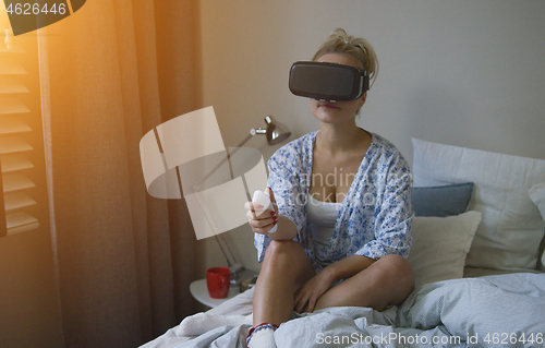 Image of Young woman in shirt and VR headset holding controller while sitting on bed