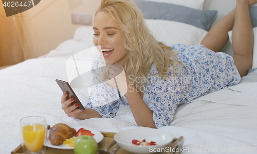 Image of Cheerful young woman browsing smartphone and laughing lying on bed