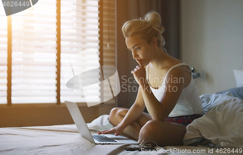 Image of Beautiful young woman in pajamas sitting on bed with laptop