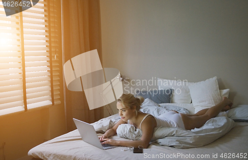 Image of Sensual young woman in white tank top and panties lying on bed near laptop