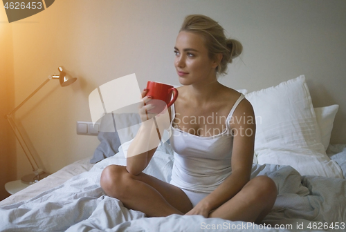 Image of Pretty woman in white tank top and panties sitting on bed and enjoying hot beverage