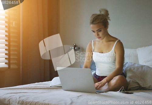 Image of Pretty young woman in shorts and tank top sitting on comfortable bed and using laptop