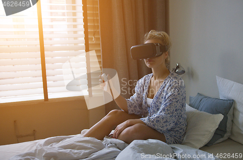 Image of Young woman in shirt sitting on comfortable bed and using VR headset with controller