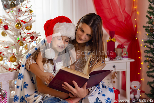 Image of Mom and daughter sit next to a Christmas tree and read a big boo