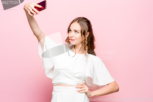 Image of Portrait of a young attractive woman making selfie photo with smartphone on a pink background