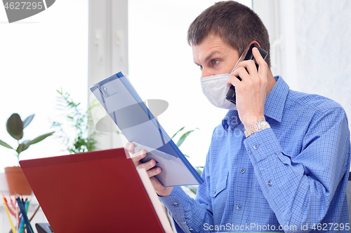 Image of A man with a medical mask on his face reads a document in the office and talks on the phone