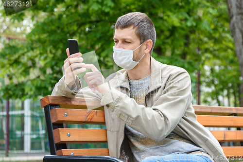 Image of A man in a medical mask with a smartphone sits on a bench in the park