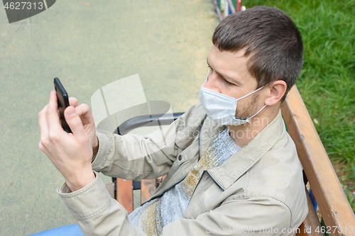 Image of A man in a protective medical mask sitting on a bench in the park communicates by cell phone