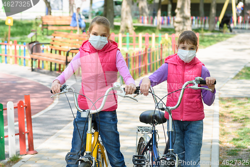Image of Two girls with medical masks on their face walk with bicycles in the playground