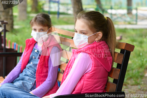 Image of Two girls in medical masks sit on a bench in a park