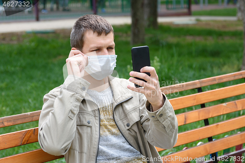 Image of A man in a medical mask sits on a bench and adjusts the mask