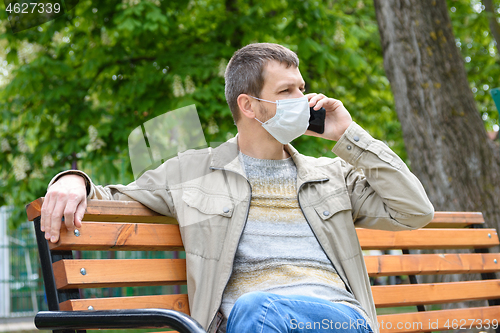 Image of A man in a protective mask walks through the park and crouches on a bench talking on the phone