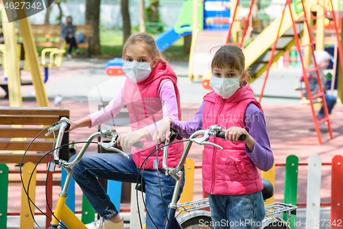 Image of Two girls in medical masks ride bicycles in the playground