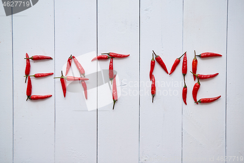 Image of A word EAT ME formed with small red chilli peppers. Placed on white wooden table