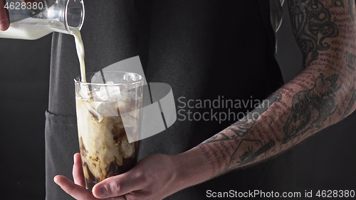 Image of A male barista with a tattoo pours fresh milk into a glass with ice and coffee.