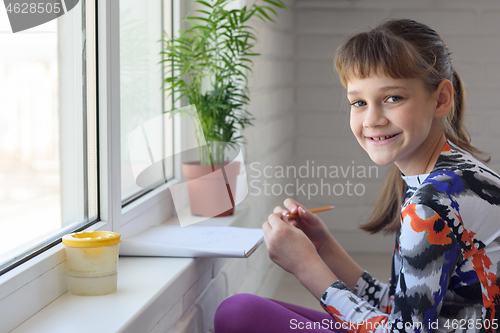 Image of Happy little girl draws a drawing while sitting on the floor near a large window