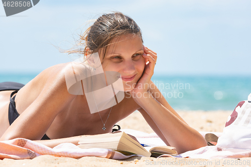 Image of Girl dreamily looks into the distance lying on a sandy beach by the sea with a book