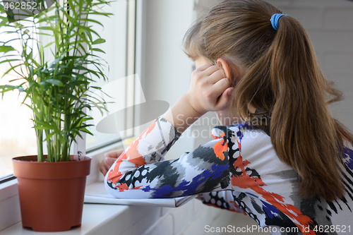 Image of A teenager looking away from the camera draws with a pencil in an album sitting on the floor in front of the window