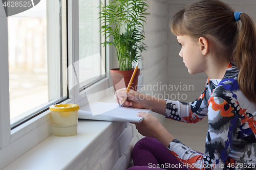 Image of Girl draws with a pencil in an album on the windowsill